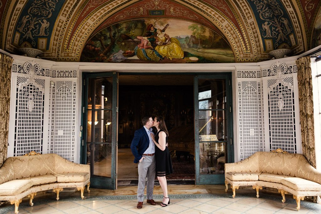 Engaged couple kisses under an ornate archway in the elegant, vintage Cuneo Mansion, surrounded by gold sofas and intricate decor