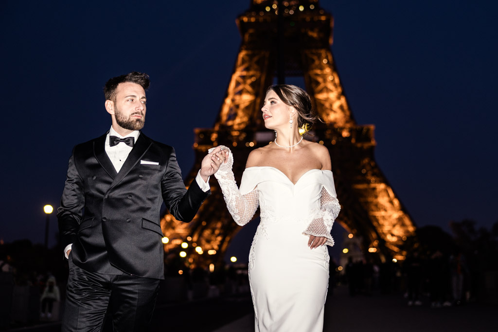 A couple in formal attire holds hands at night in front of the illuminated Eiffel Tower, capturing a magical Eiffel Tower wedding moment.