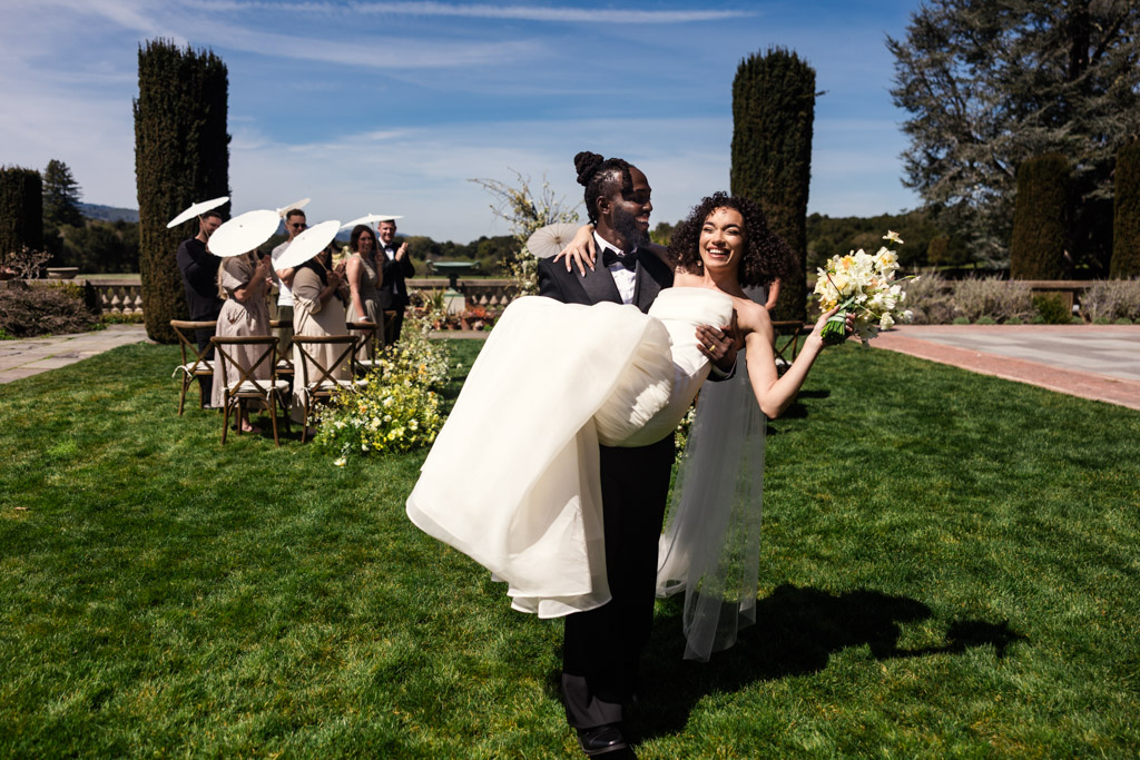 Happy bride and groom exit their garden wedding ceremony at Filoli Historic House and Gardens as guests smile at them holding parasols in the California sunshine