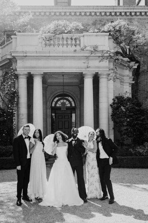 Black and white photo of wedding party in formal attire stands smiling with parasols in front of Filoli Historic House