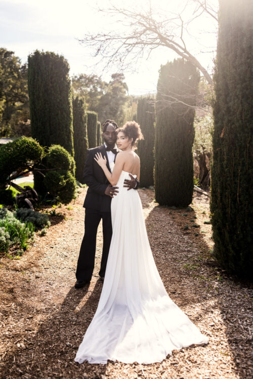 Portrait of bride and groom stand together on a sunlit garden path lined with tall hedges and trees at Filoli Historic House