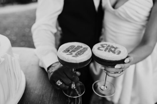 Bride and groom holding custom cocktails with decorative words written in foam, standing next to a wedding cake at Filoli Historic House & Gardens