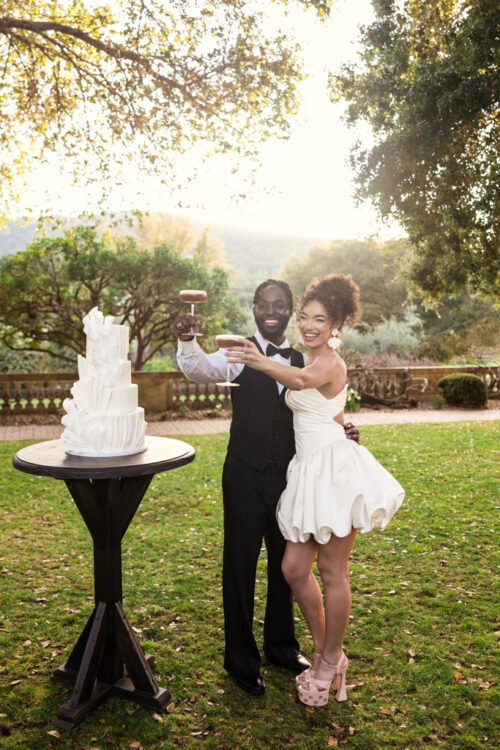 Newlywed couple in formal attire raise a toast by a white wedding cake outdoors on a sunny day at Filoli Historic House & Gardens in California