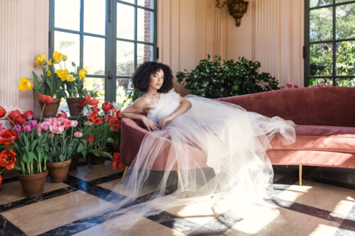Bride in a white tulle gown lounges on a pink sofa, surrounded by colorful potted flowers in a sunlit room at Filoli Historic House & Gardens