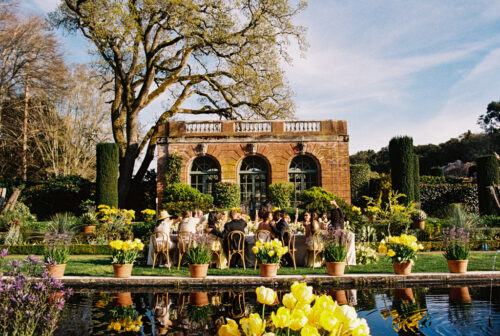 Newlyweds and their guests dine outdoors at a long table in a lush garden near a historic brick building during Filoli Historic House & Gardens wedding reception