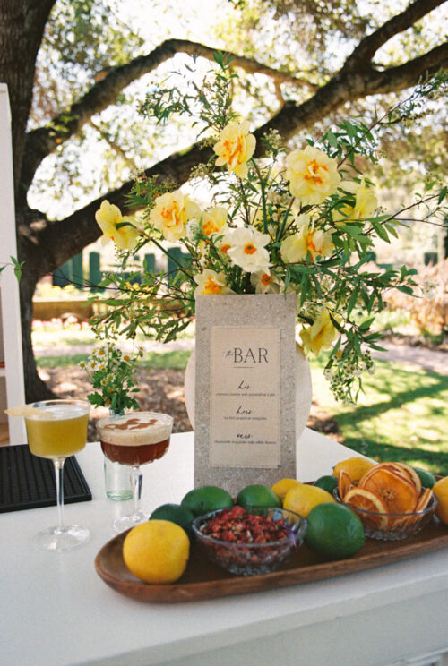 Outdoor bar setup for wedding reception at Filoli Historic House & Gardens featuring yellow flowers, a cocktail menu, fresh citrus fruits, and a bowl of pomegranate seeds