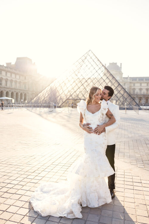 A bride and groom pose in front of the Louvre Pyramid during sunset, capturing the romance of a Paris elopement.