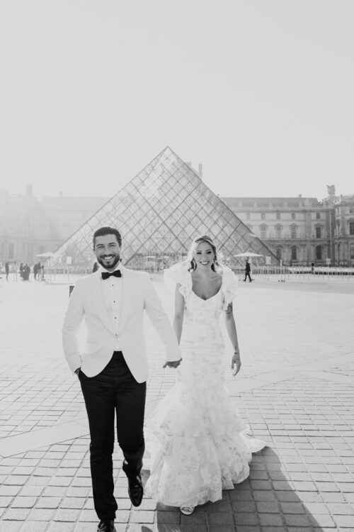 A bride and groom smile, walking hand in hand outside the Louvre Pyramid, capturing the magic of their Paris elopement.