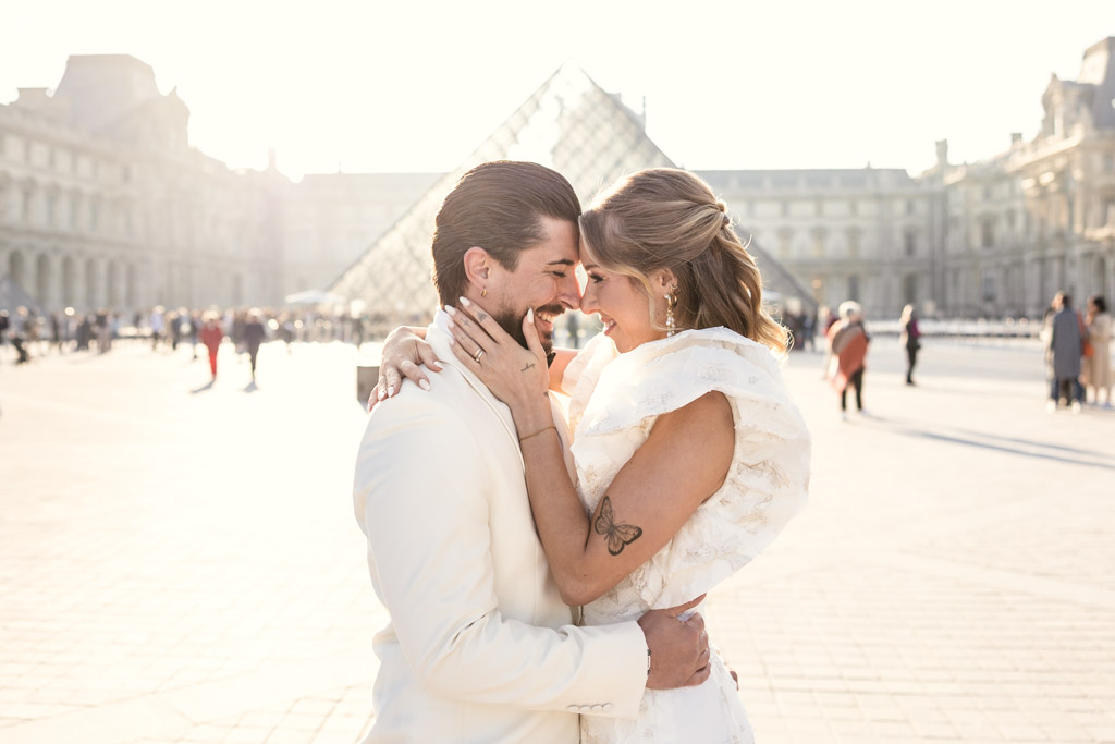 A couple in wedding attire embraces and smiles in front of the Louvre Pyramid, capturing the magic of a Paris elopement.