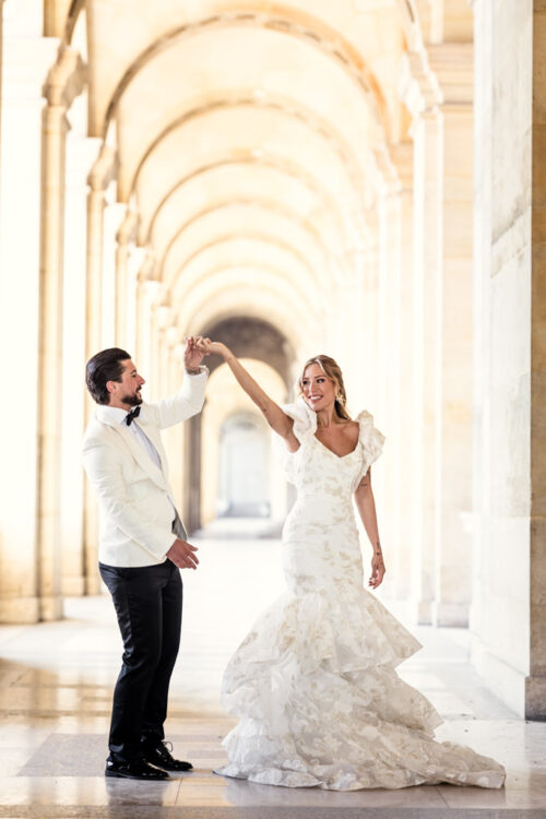 A bride and groom dance in a sunlit, arched hallway during their Paris elopement, both smiling and dressed in formal wedding attire.