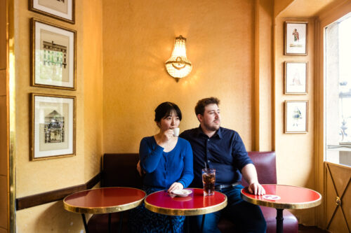 Two people sit at red tables in a cozy cafe; the woman, perhaps celebrating a Paris engagement, sips from a cup while the man looks away.