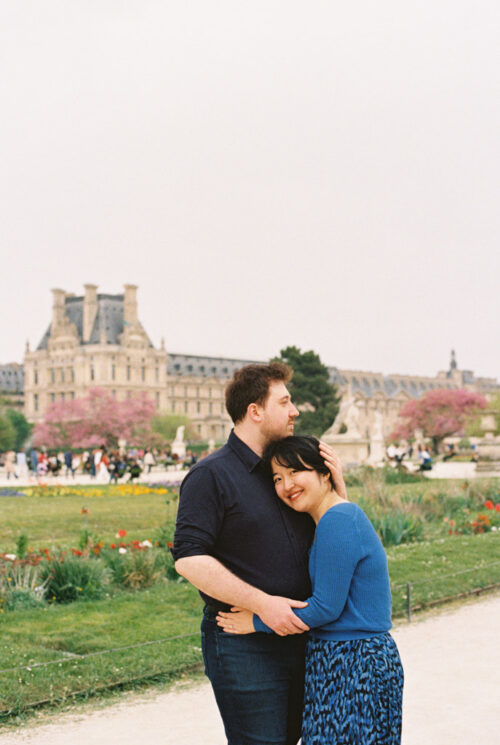 A couple embraces and smiles in a garden with the Louvre Museum in the background, capturing the joy of their Paris cafe engagement.