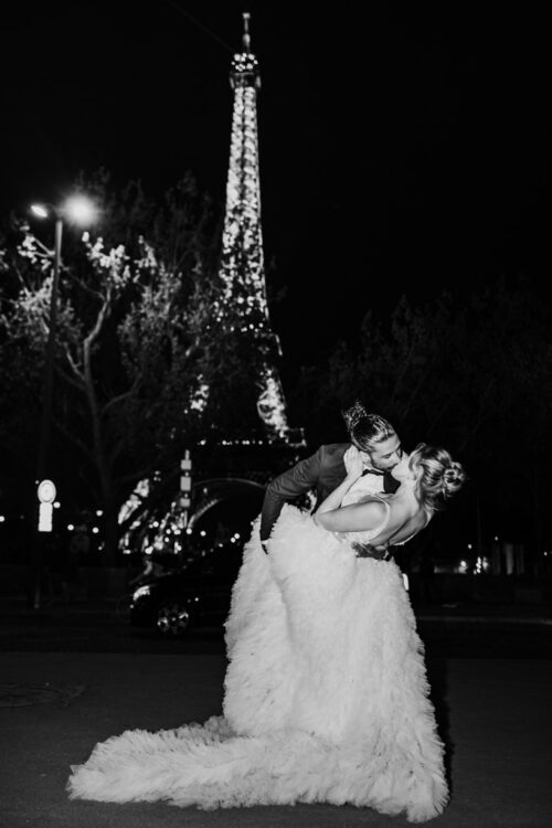 A couple shares a kiss at night in front of the illuminated Eiffel Tower; the bride wears a fluffy gown, capturing the magic of a Paris wedding.