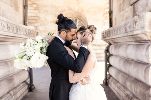 Bride and groom embracing closely, holding a bouquet, standing between stone walls—capturing the romance of a Paris wedding.
