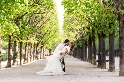 A groom dips his bride for a kiss under a canopy of green trees in a sunlit outdoor walkway, capturing the romance of a Paris elopement.