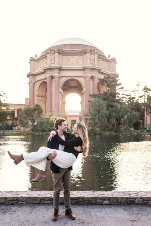 Man holds his fiancée in his arms by a pond, with the Palace of Fine Arts in the background during San Francisco engagement session