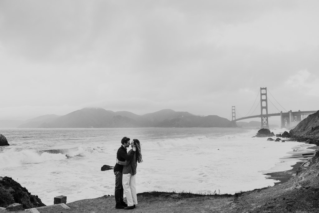 Moody and romantic photo of couple embracing on San Francisco coast at Marshall's Beach with Golden Gate Bridge in the background and fog rolling in