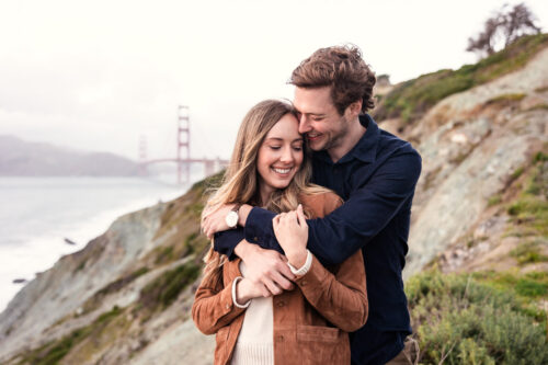 Engaged couple embraces and smiles on a hillside at Marshall's Beach near San Francisco with the Golden Gate Bridge in the background
