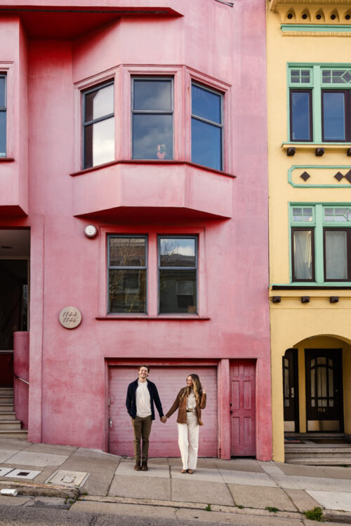 Couple holds hands standing in front of a pink apartment building next to a yellow building during engagement session in Nob Hill neighborhood in San Francisco