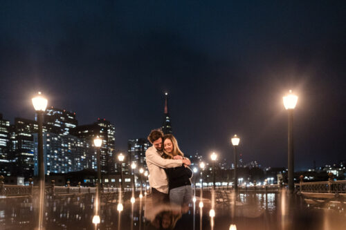 Creative photo of engaged couple holding each other on a San Francisco city pier at night, surrounded by glowing streetlights and towering skyscrapers