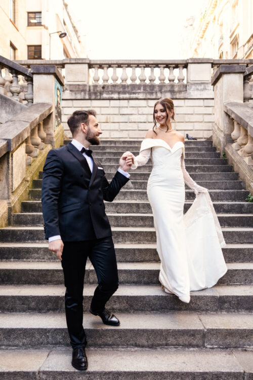 A groom helps his bride in a white dress walk down outdoor stone steps, both smiling, as the Eiffel Tower wedding venue stands majestically in the background.