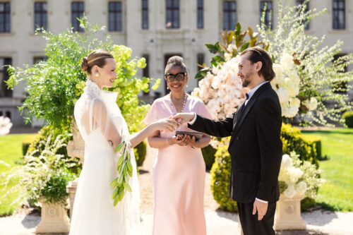 A bride and groom exchange rings outside Chateau Challain in Loire Valley, France during their wedding ceremony, with an officiant smiling behind them.