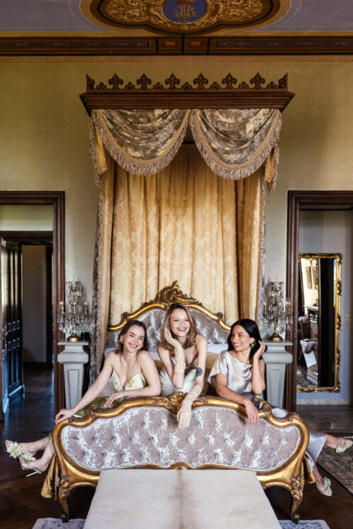Bride and bridesmaids sit and pose on a gold and cream canopy bed in an ornate room inside Chateau Challain, Loire Valley, France