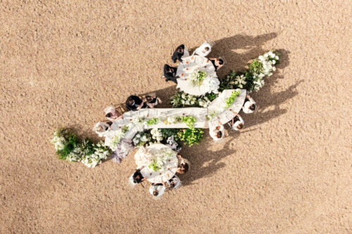 Aerial view of people dining at decorated round and curved tables on a sandy surface, evoking the elegance of a French castle wedding.