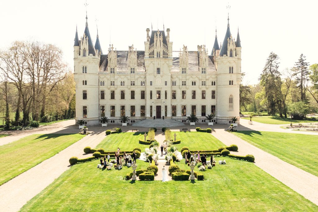 Large white French castle with towers, manicured gardens, and people gathered on the lawn for a wedding under bright daylight.