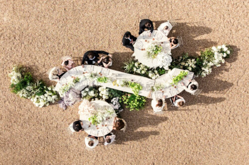 Aerial view of people dining at decorated round and curved tables on a sandy surface, evoking the elegance of a French castle wedding.