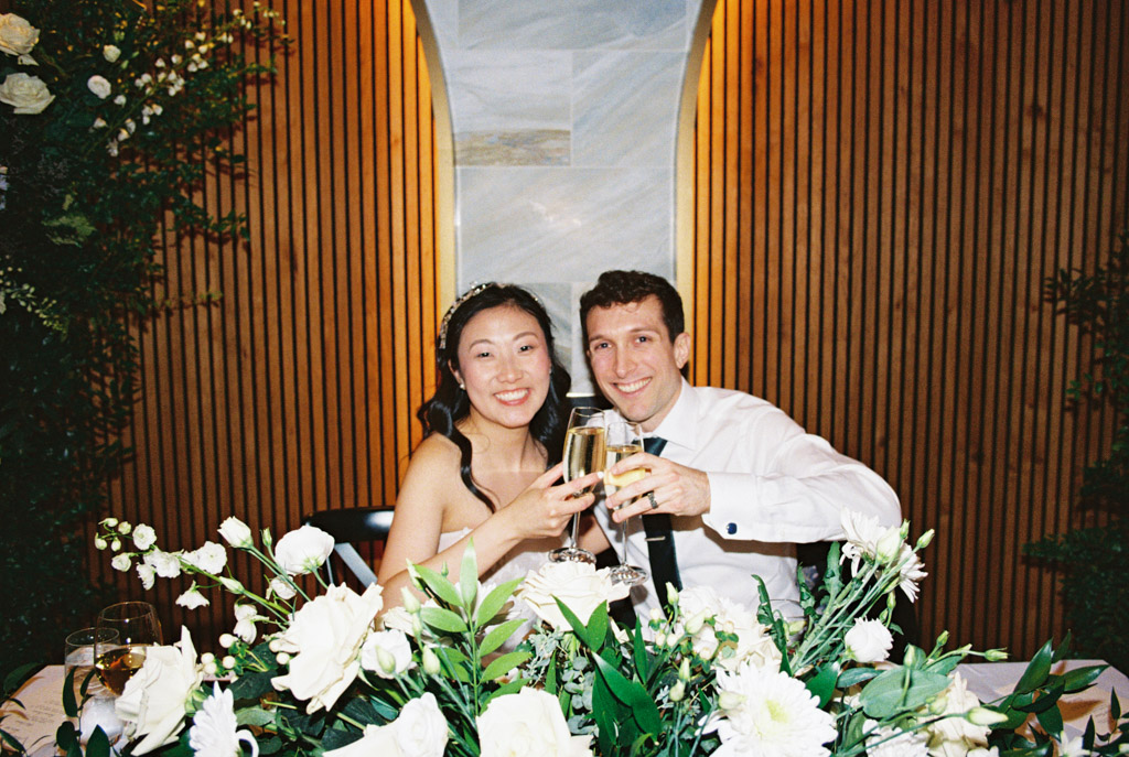 happy bride and groom in wedding attire toast with champagne behind a table of white flowers during their Chicago Winery wedding reception