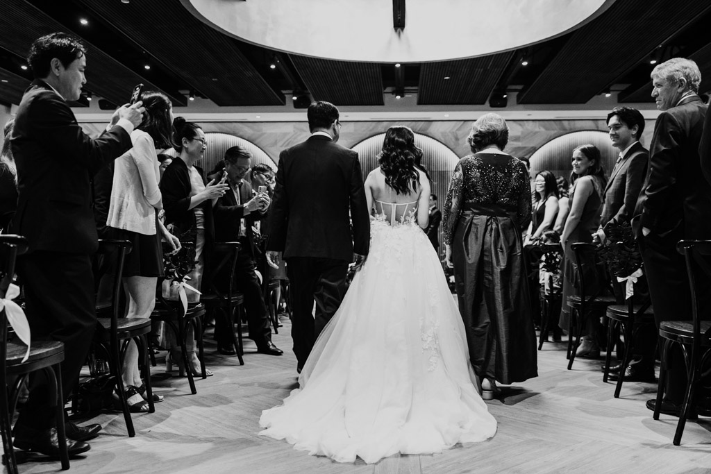 Black and white photo of bride in a gown walking down the aisle with her parents, surrounded by seated guests at Chicago Winery wedding ceremony
