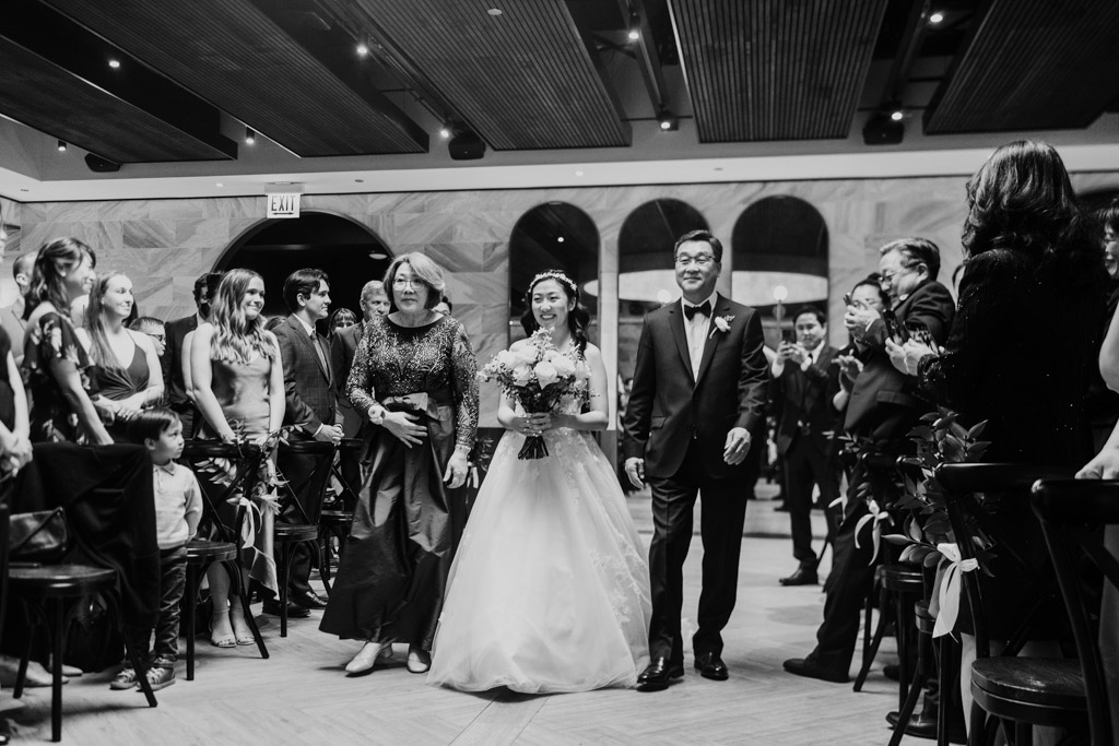 Bride walks down the aisle with her parents during her wedding ceremony at Chicago Winery, surrounded by smiling guests