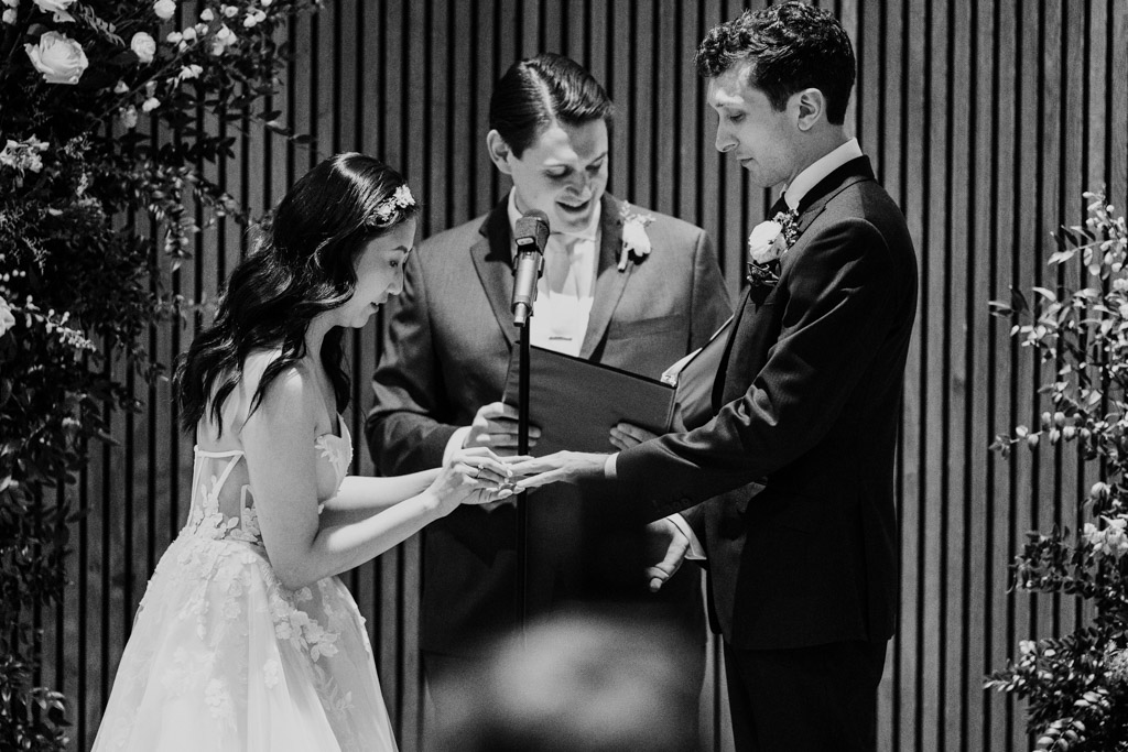Black and white photo of bride placing ring on groom's finger during their wedding ceremony at Chicago Winery, with an officiant standing behind them