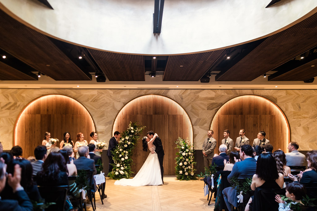 Bride and groom kiss during their wedding ceremony at Chicago Winery, surrounded by their wedding party and guests