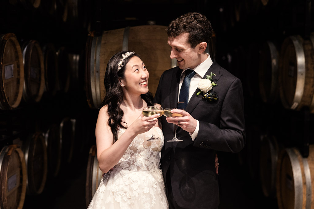 Bride and groom smiling and toasting with champagne in a wine cellar surrounded by barrels at Chicago Winery