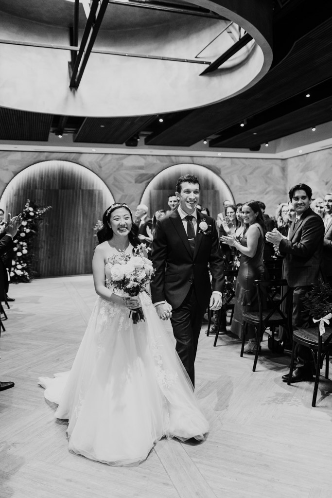 Black and white photo of bride and groom walking down the aisle, smiling as guests applaud during wedding ceremony at Chicago Winery