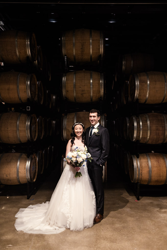 Portrait of bride and groom smiling, standing in front of stacked wine barrels in the Chicago Winery barrel room