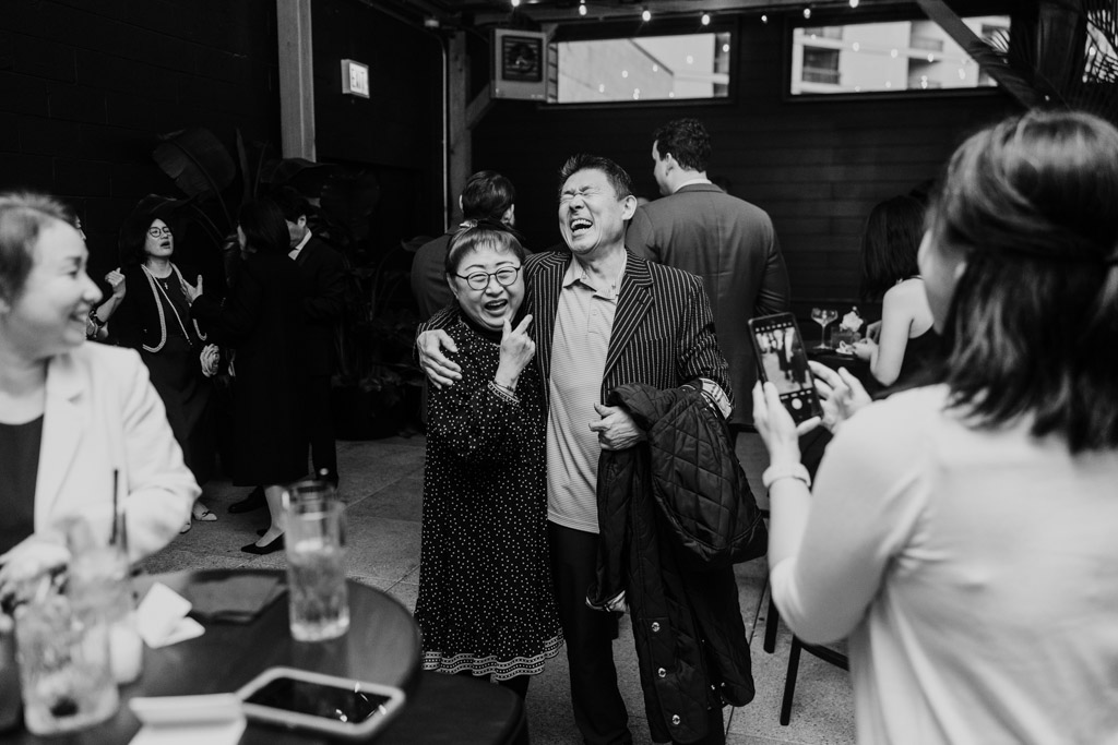 Black and white photo of two guests smiling and laughing as others take photos of them at Chicago Winery wedding reception