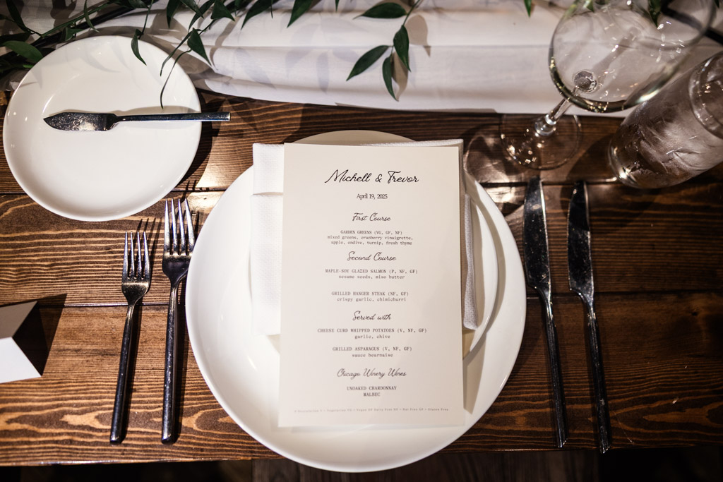 A wedding dinner menu on a white plate with cutlery, wine glasses, and greenery on a wooden table for Chicago Winery wedding reception