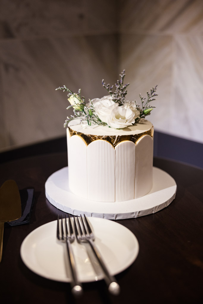 Elegant white wedding cake with gold accents, topped with white flowers and greenery for wedding reception at Chicago Winery on a table with plates and forks