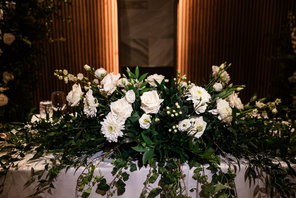 A floral arrangement with white roses and greenery decorates the head table with a white tablecloth for Chicago Winery wedding reception