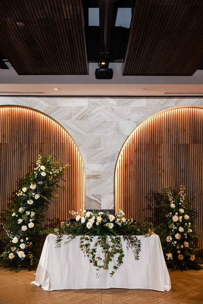 Head table with a white cloth and floral arrangements in front of a wooden wall with two large, arched insets inside Chicago Winery wedding reception space