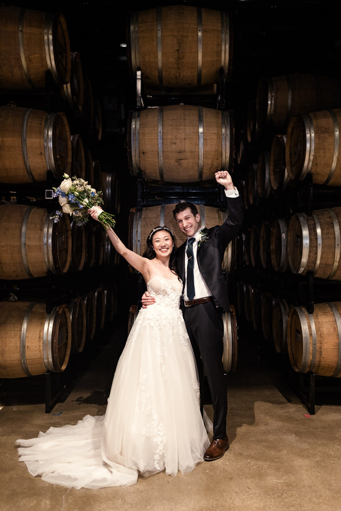 Bride and groom raise their arms in celebration in front of stacked wine barrels at Chicago Winery