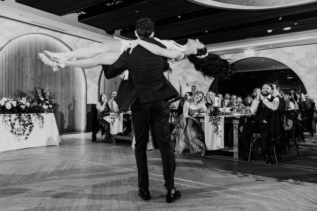 Black and white photo of groom lifting the bride during their first dance as guests watch and smile at Chicago Winery