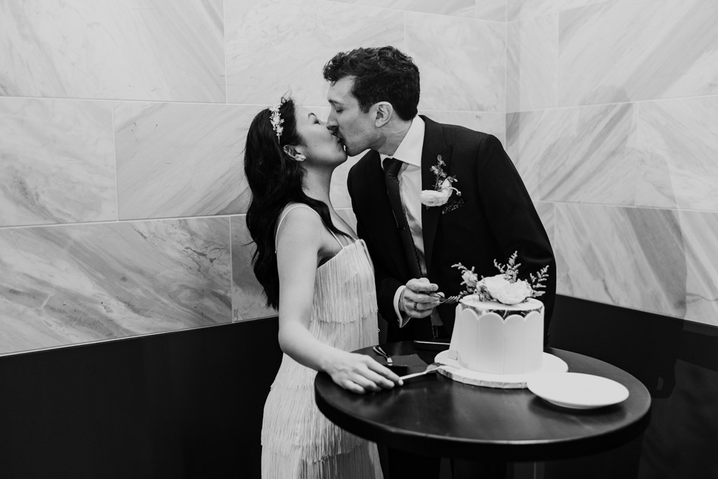 Black and white photo of bride and groom kissing while cutting their cake during wedding reception at Chicago Winery