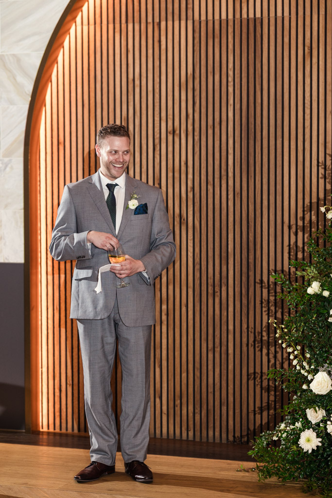 Best Man in a gray suit holding a drink, smiling, stands by a wooden wall with flowers on the right during Chicago Winery wedding reception