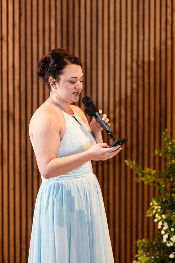 Maid of Honor in a light blue dress speaks into a microphone while reading from her phone in front of a wooden wall during Chicago Winery wedding reception