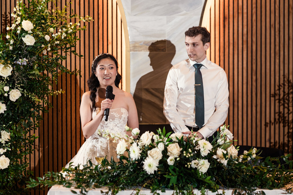 Bride speaks into a microphone beside the groom, standing behind a floral arrangement during their Chicago Winery wedding reception