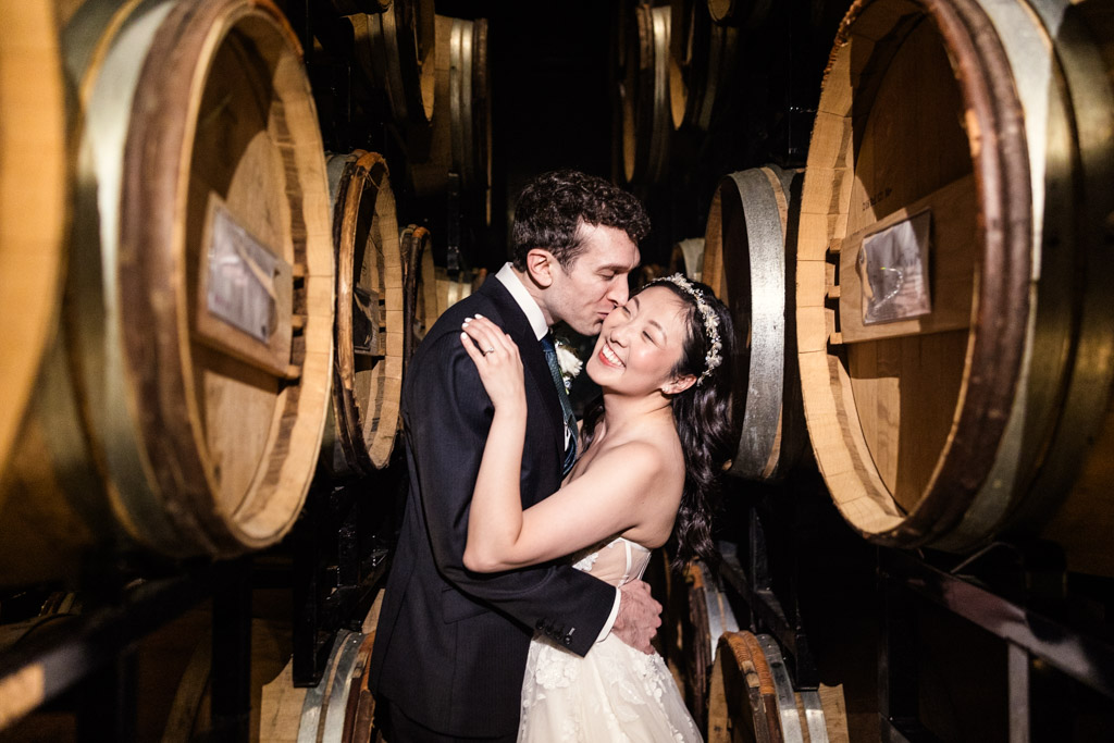 Groom kisses bride's cheek while they stand between stacks of large wine barrels in the Chicago Winery barrel room